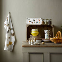Kitchen setting with a towel featuring chicken illustrations, a tray with rooster design, and various kitchenware on a wooden shelf.